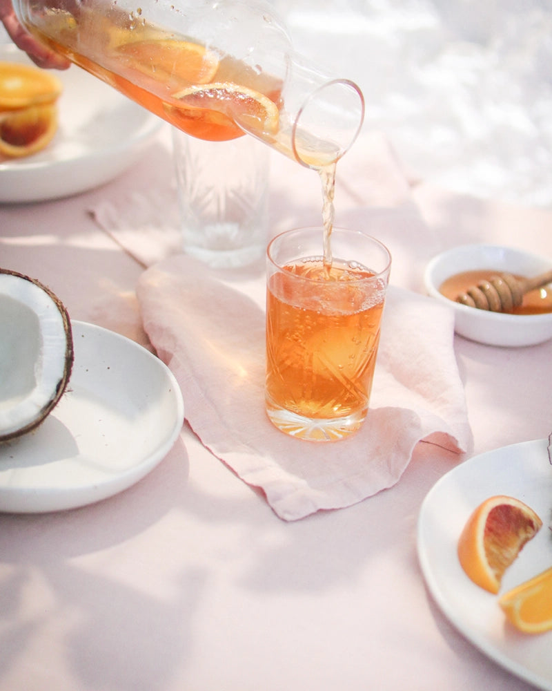 Pouring a refreshing glass of Coconut Orange Iced Tea on a sunlit table, accompanied by sliced oranges and a coconut shell.