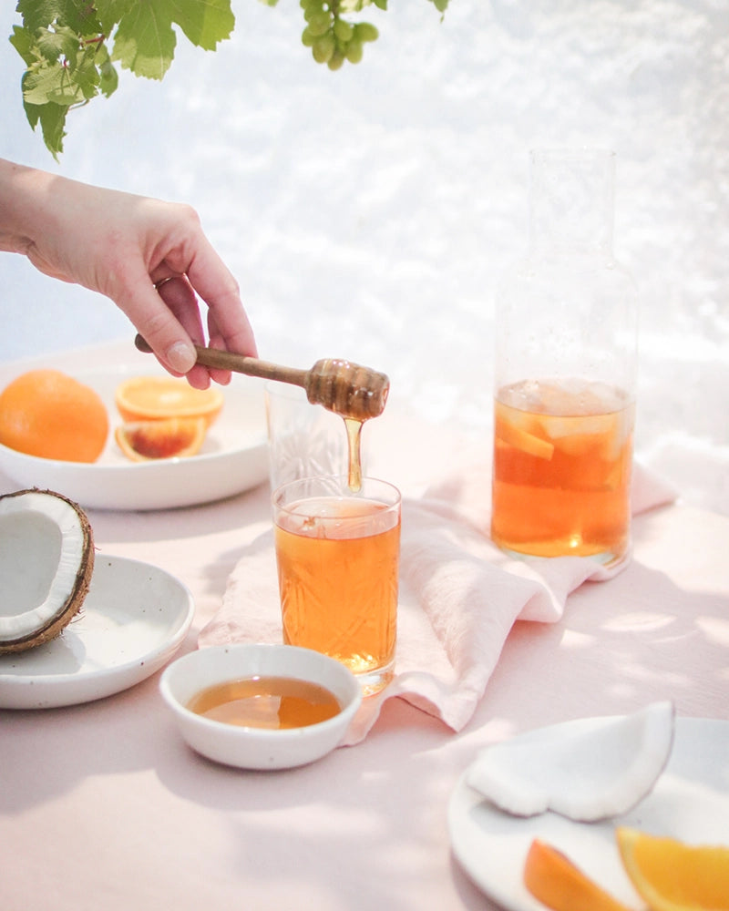 Adding honey to a glass of Coconut Orange Iced Tea, with fresh oranges on a pastel cloth background