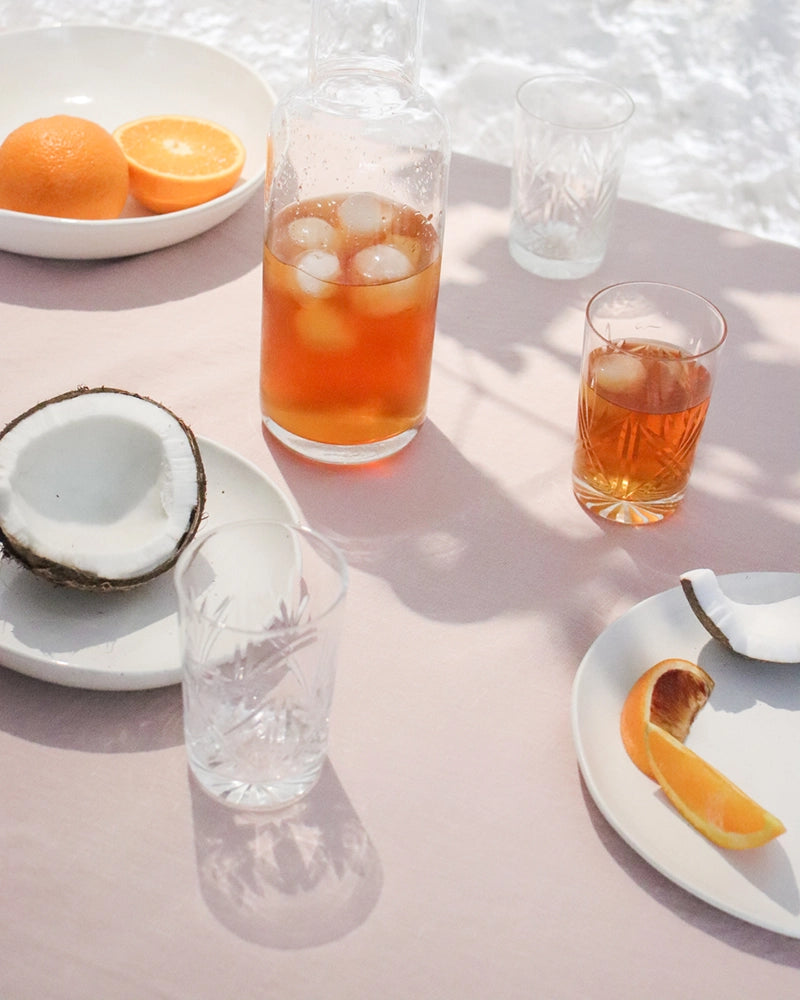 A serene outdoor setting featuring a glass of Coconut Orange Iced Tea, a coconut shell, and whole oranges on a pastel tablecloth