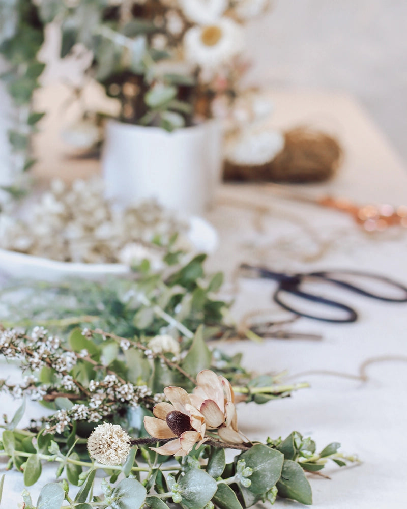 Crafting materials for a native foliage garland, including twine, shears, and a variety of greenery on a linen backdrop.