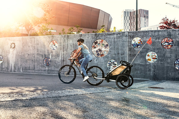Mutter fährt Fahrrad und zieht einen thule Multisporttrailer Lite durch moderne Stadtkulisse