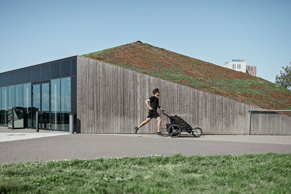 Architektenhaus mit Holzverkleidung in Skandinavien. Im Vordergrund joggt ein in schwarz gekleideter Vater mit seinem Multisport-Trailer von Thule. Modell Sport 2, Black
