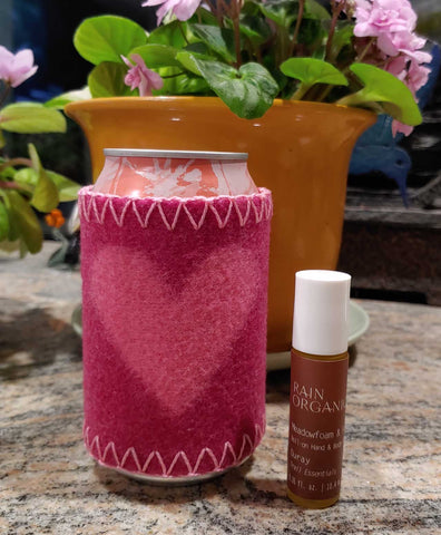 pink Merino wool sleeve coozie for aluminum cans with pink heart and red Ouray rollerball for chapped hands with blooming light pink African violet blooming in background