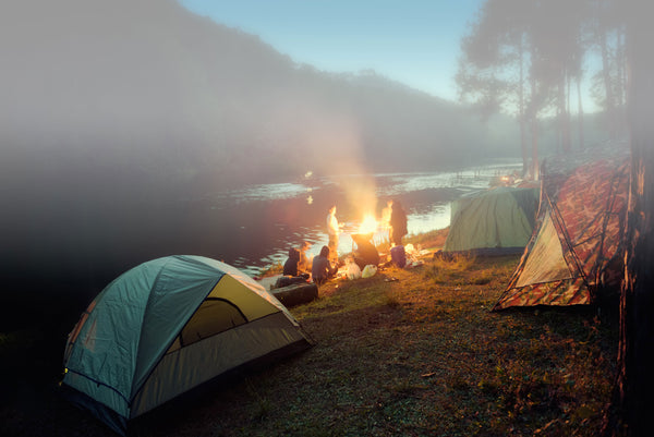 people gathered around a campfire next to a river with tents set up
