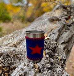 stainless steel tumbler wrapped in navy blue wool sleeve with orange star on it sitting on a stump with fall colored forest in background