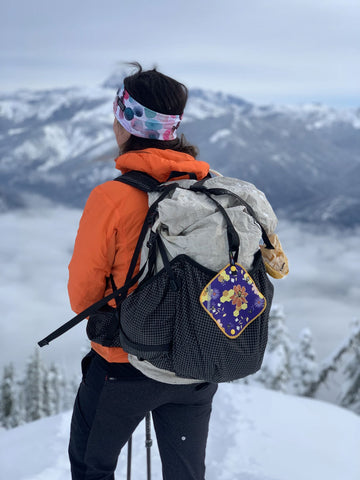 woman backpacking in winter wearing an orange coat standing atop a snow covered mountain vista overlooking soft curvaceous mountain peaks with a beehive Kula Cloth attached to her backpack