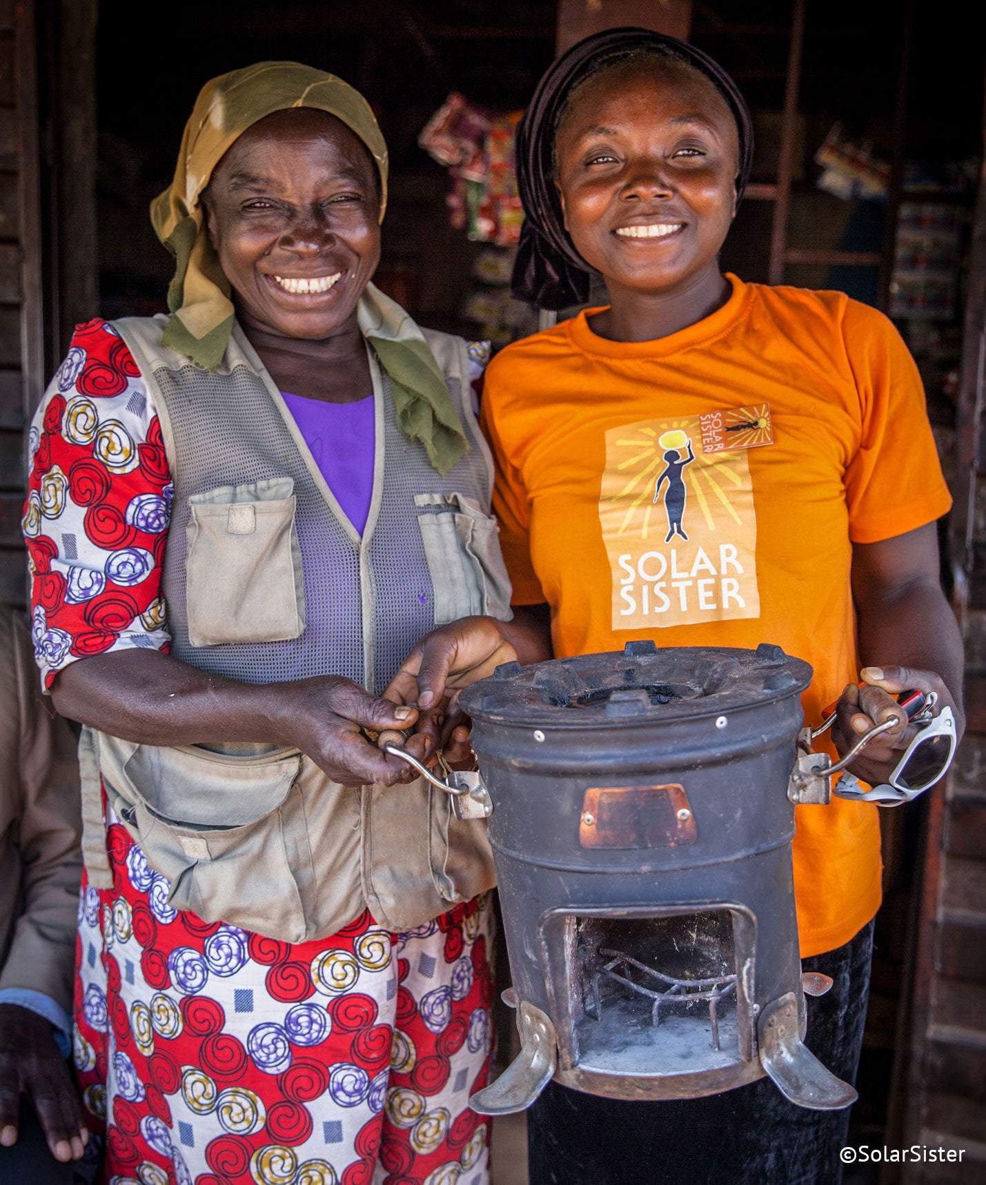 Two women stand, smiling at the camera, holding a fire stove. The woman on the right wears an orange Solar Sister t-shirt.