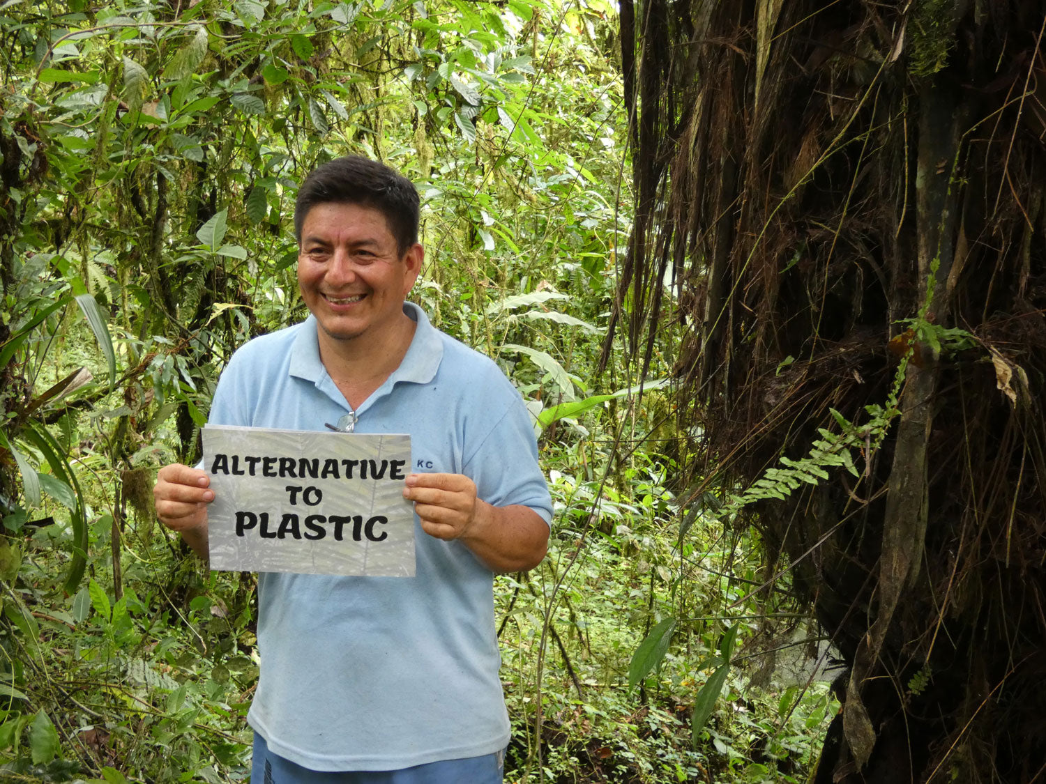 Man stands in the Ecuador rainforest, smiling, holding a sign which reads 'Alternative to plastic'