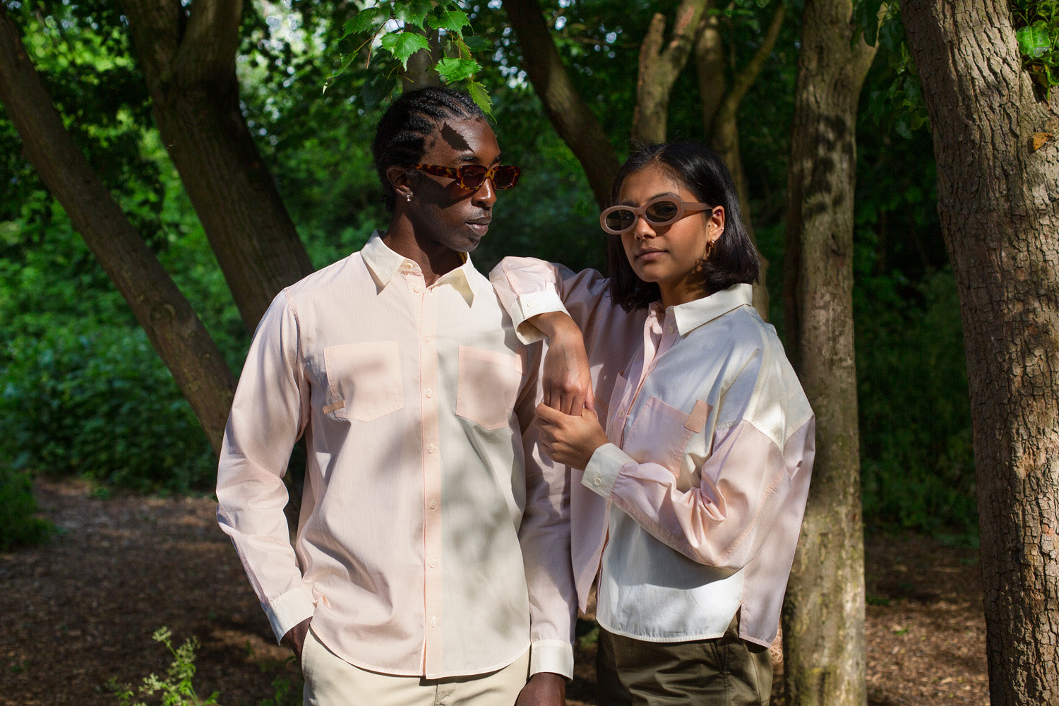 Two models stand side by side, under tree cover. He looks at her, whilst she has her elbow on his shoulder. They both wear pastel orange and yellow shirts and sunglasses. Saywood sustainable menswear and womenswear.