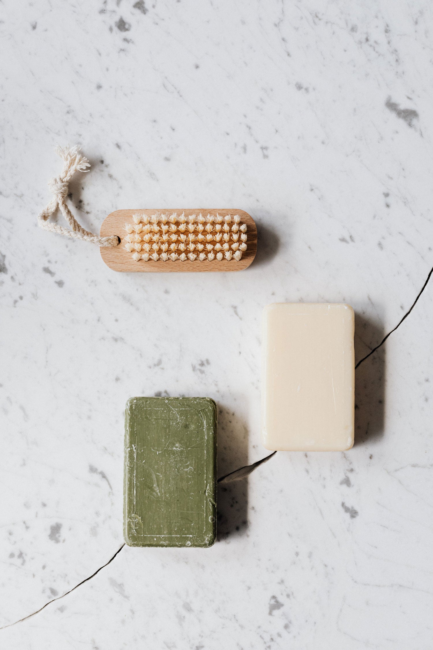 Image shows two bars of soap, one in green and one in cream, laying on a grey white marbled surface. A scrubbing brush lies next to them.