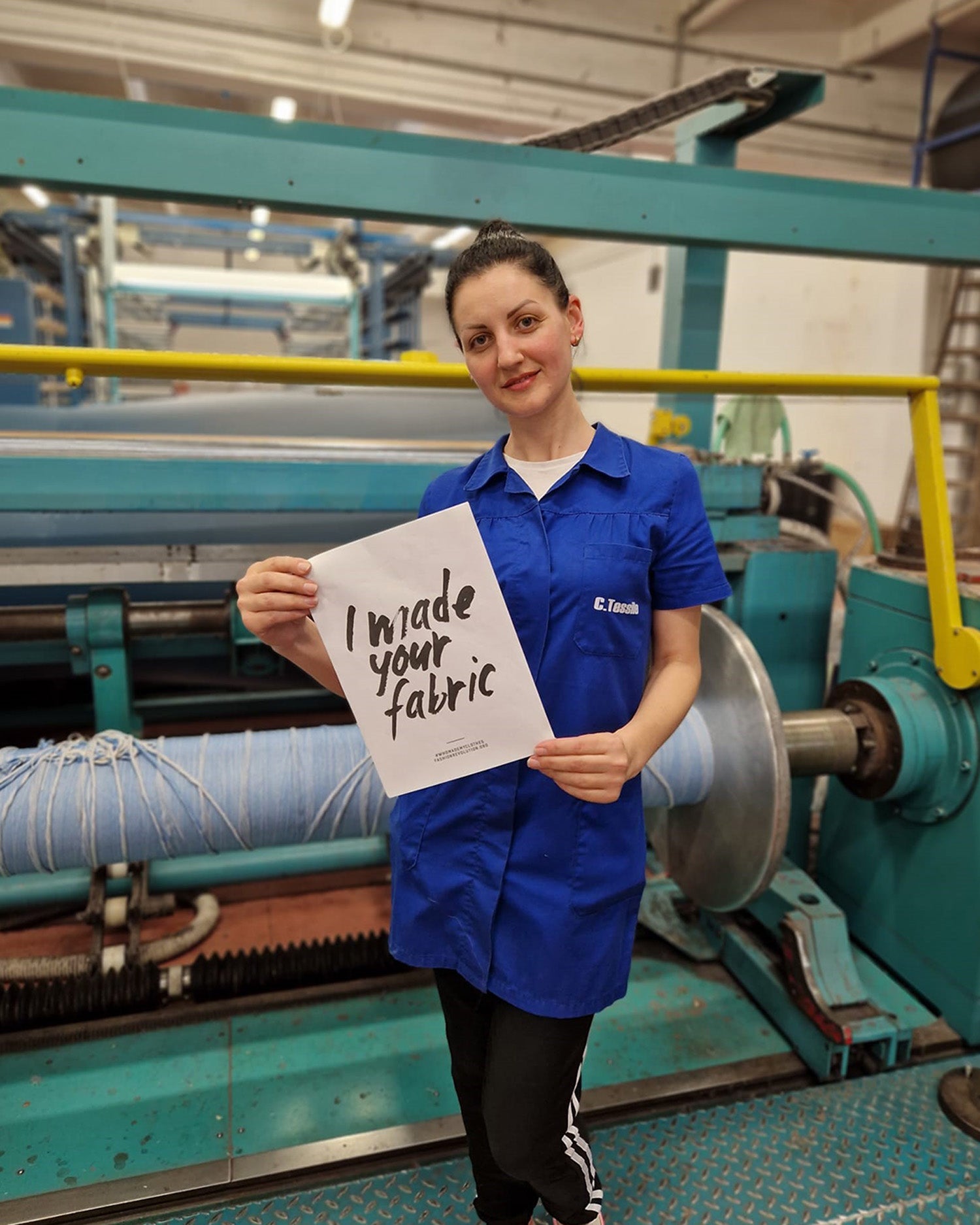 Fabric maker and warper, Guilia, stands in front of a fabric machine in the fabric mill, Canclini in Italy, wearing a blue coat, holding a sign saying 'I made your fabric'.