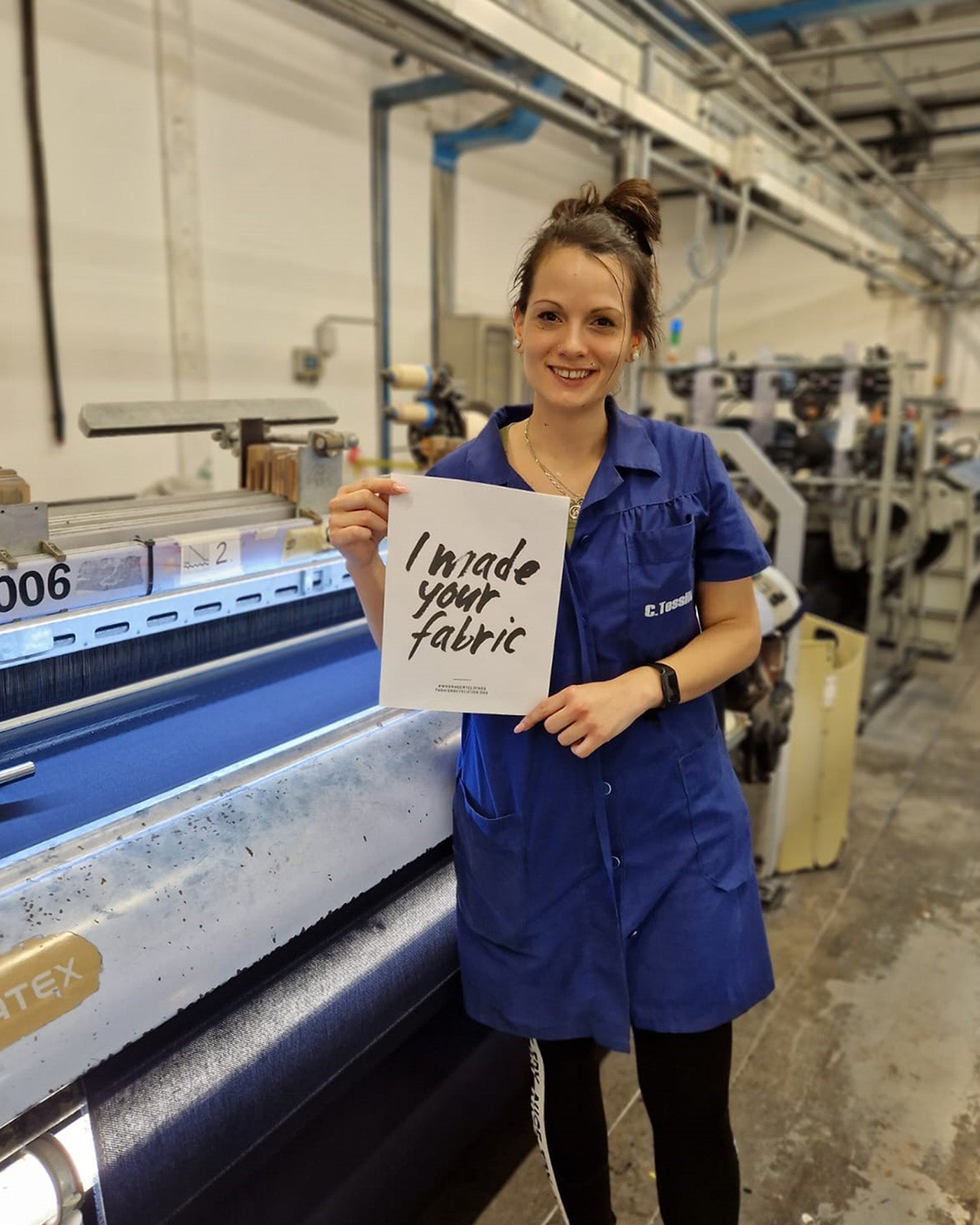 Fabric maker and weaver, Susanna, stands in front of a weaving machine in the fabric mill, Canclini in Italy, wearing a blue coat, holding a sign saying 'I made your fabric'.