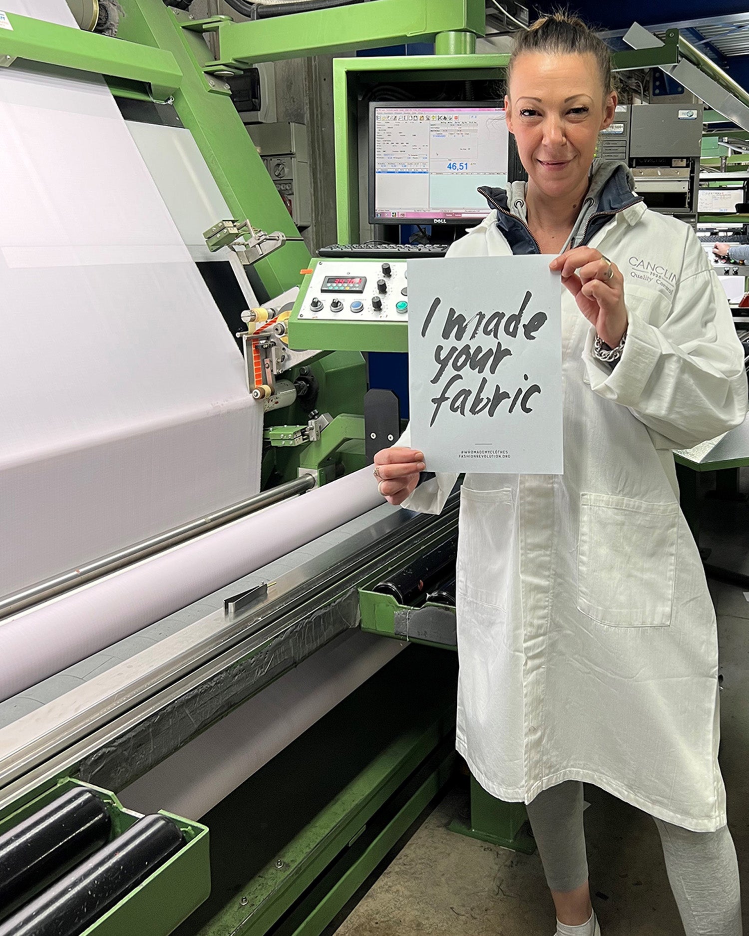 Fabric maker and quality control specialist, Daneilla, stands in front of a fabric machine in the fabric mill, Canclini in Italy, wearing a white coat, holding a sign saying 'I made your fabric'.