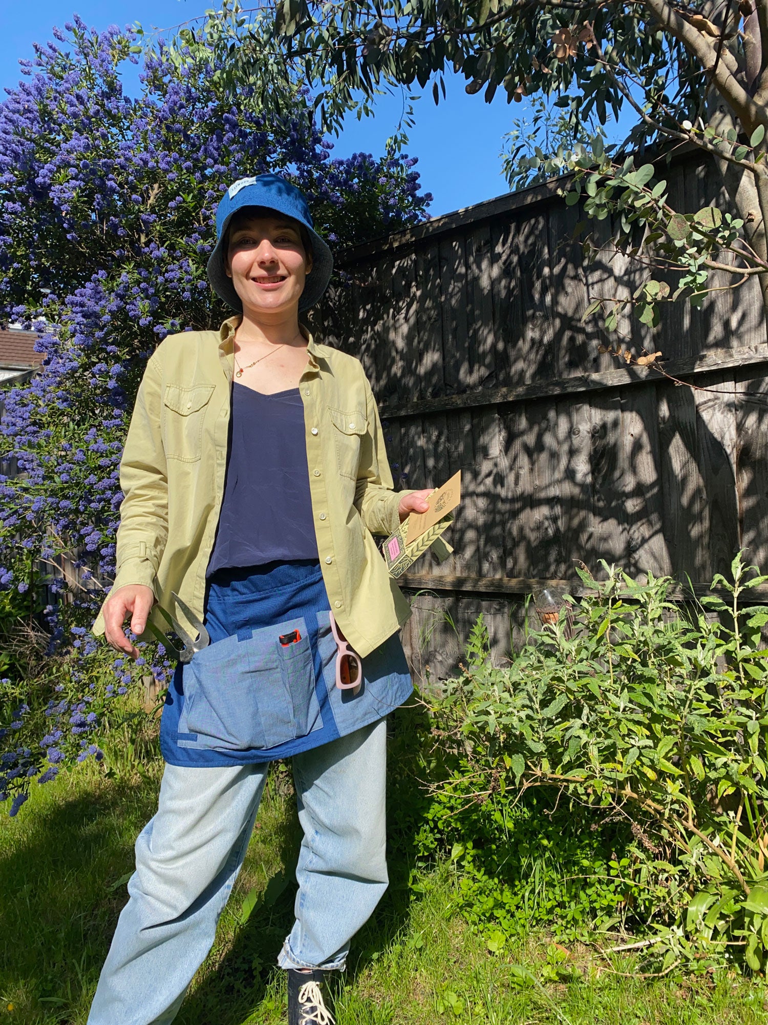 Saywood founder Harriet in the garden wearing the Garden Tool Belt around her waist in Japanese Denim. Worn with the Zadie Boyfriend Shirt in olive, jeans, and the Japanese Denim Reversible Bucket Hat