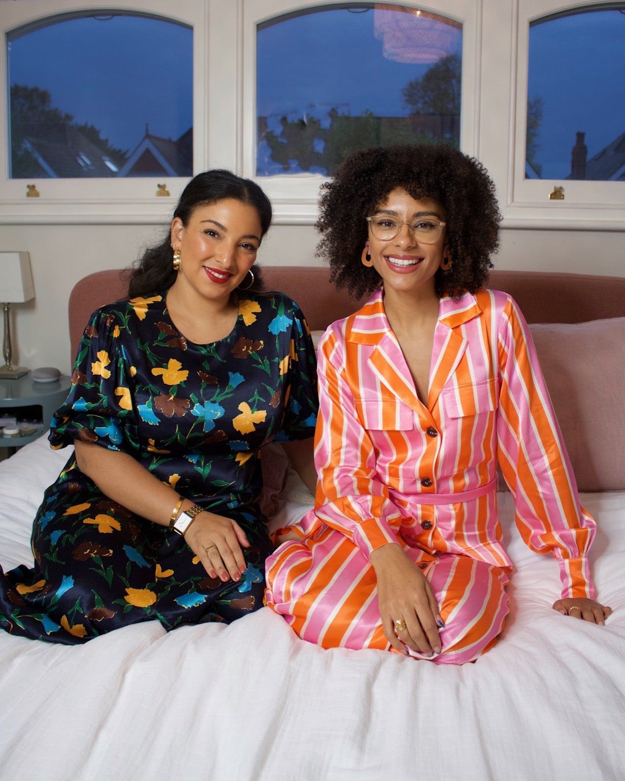 Sustainably Influenced Podcast co-founders and influencers Bianca F Foley and Charlotte Williams.  Bianca wears a black dress with a blue, ocre and chocolate print. Charlotte wears a pink an orange bold stripe pyjama set. They both are sitting on a bed.
