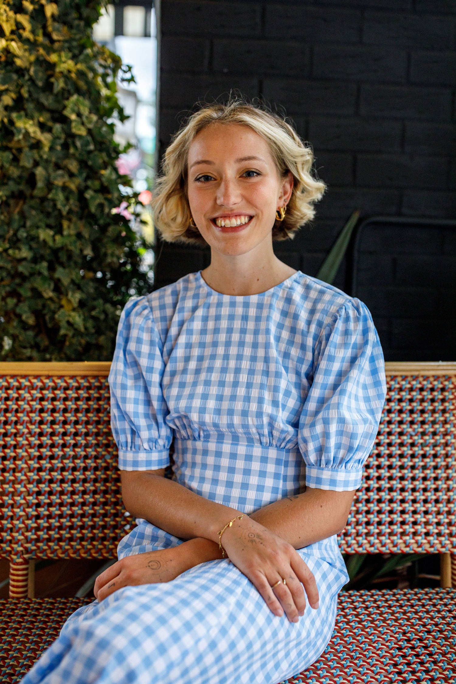 Lofte founder Daisy Harvey sits with her arms softly folded in her lap. She wears a blue white gingham dress, and she smiles.
