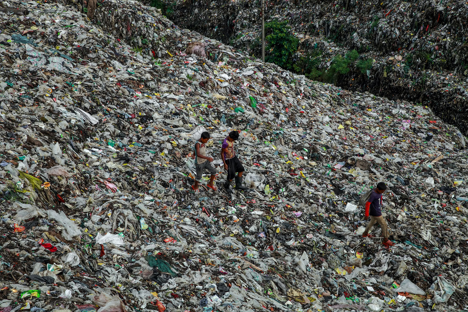 Landfill site with too much waste, from plastic to food to general litter. Three people are walking over the landfill waste pile.