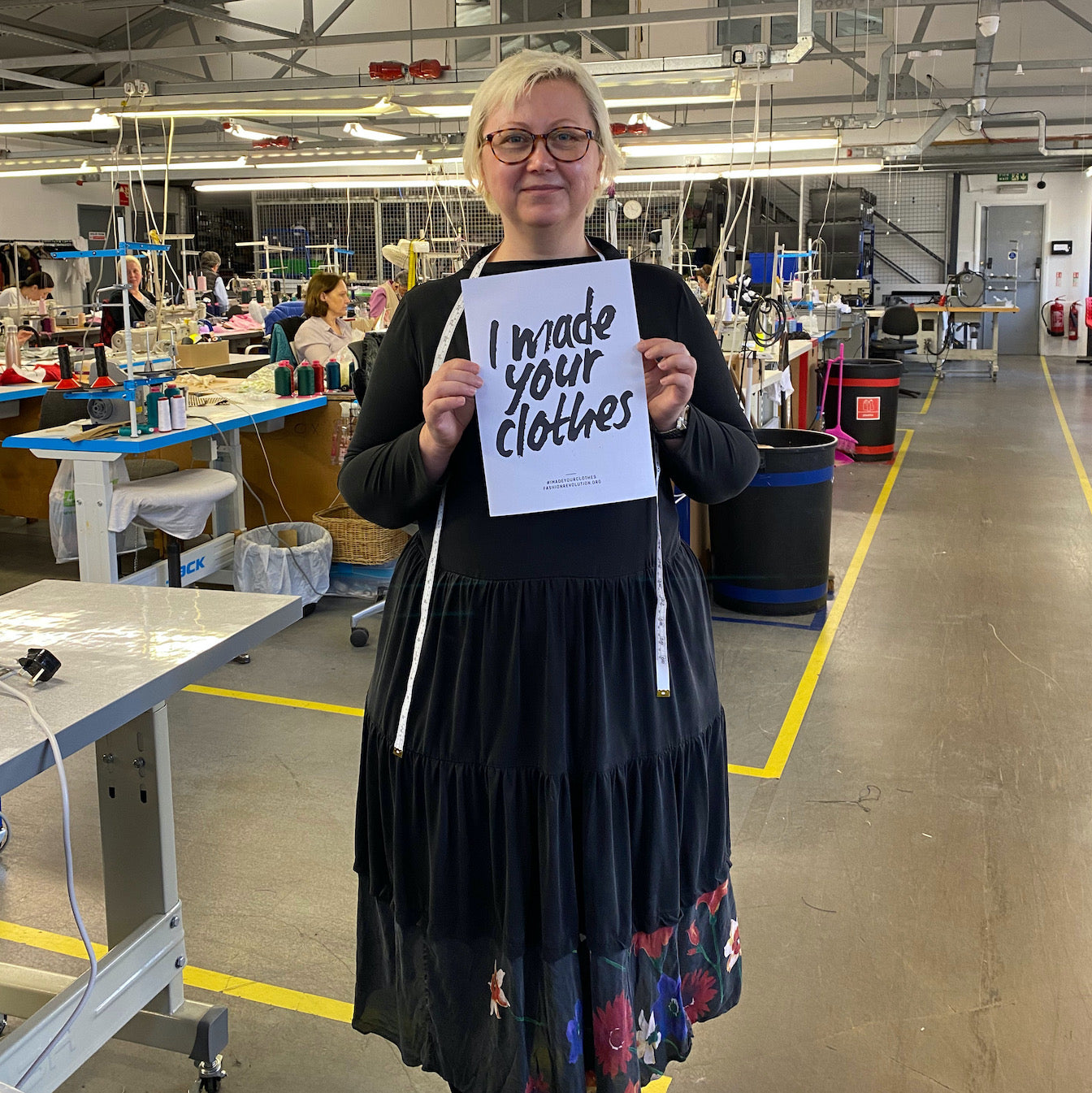 Image shows a woman and garment worker standing, smiling, and holding a sign with the text 'I made your clothes' for Fashion Revolution Week. This is Viola, a presser from Saywood's ethical London manufacturer, Apparel Tasker.