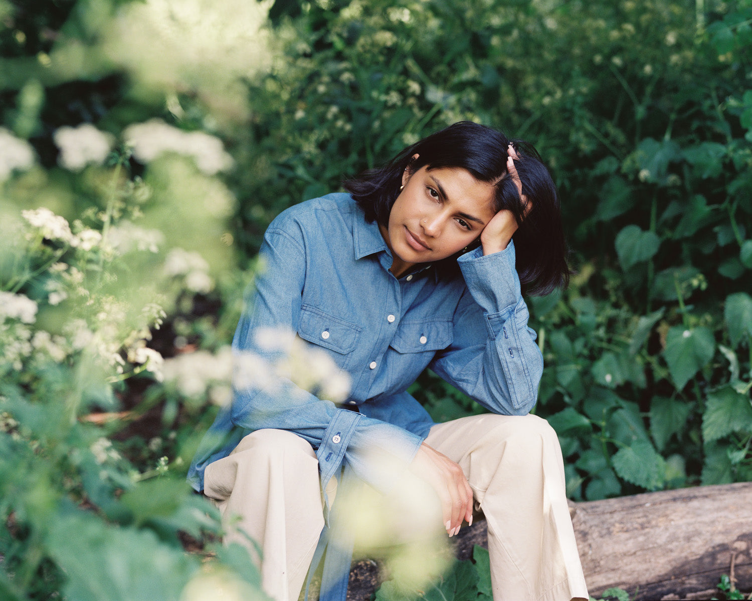 Model sits on a tree log surrounded by nature and echinacea flowers, wearing Saywood's Zadie Japanese Denim Oversized Shirt with beige trousers.
