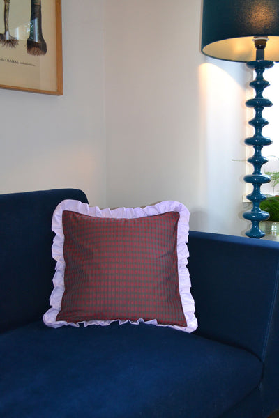 Home Textiles Saywood Ruffle Cushion from deadstock, in red check with a lilac frill edge. The cushion is placed on a navy velvet sofa, with a blue tall lamp and picture from visible