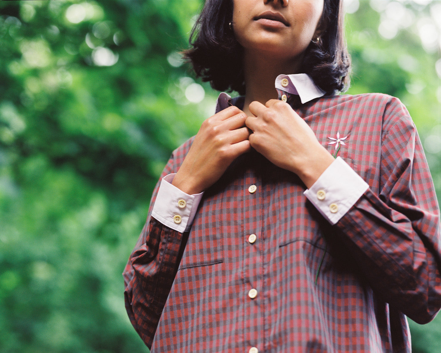 Close up of Etta shirtdress in red check with lilac collar and cuffs. Model's hands are touching to the neck of the shirt. Green foliage can be seen behind model.