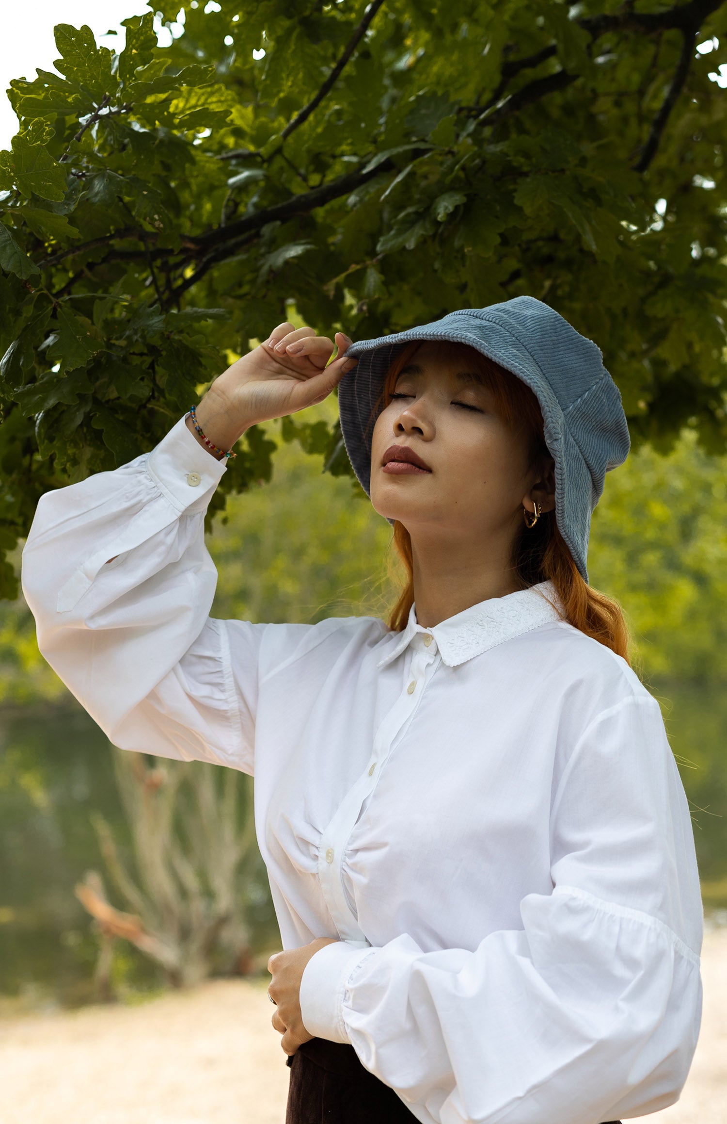 Model wears Saywood's Edi Volume Sleeve Shirt in white Supima cotton and bamboo. She has one hand across her waist, and the other hand holding the tip of a blue cord bucket hat on her head.