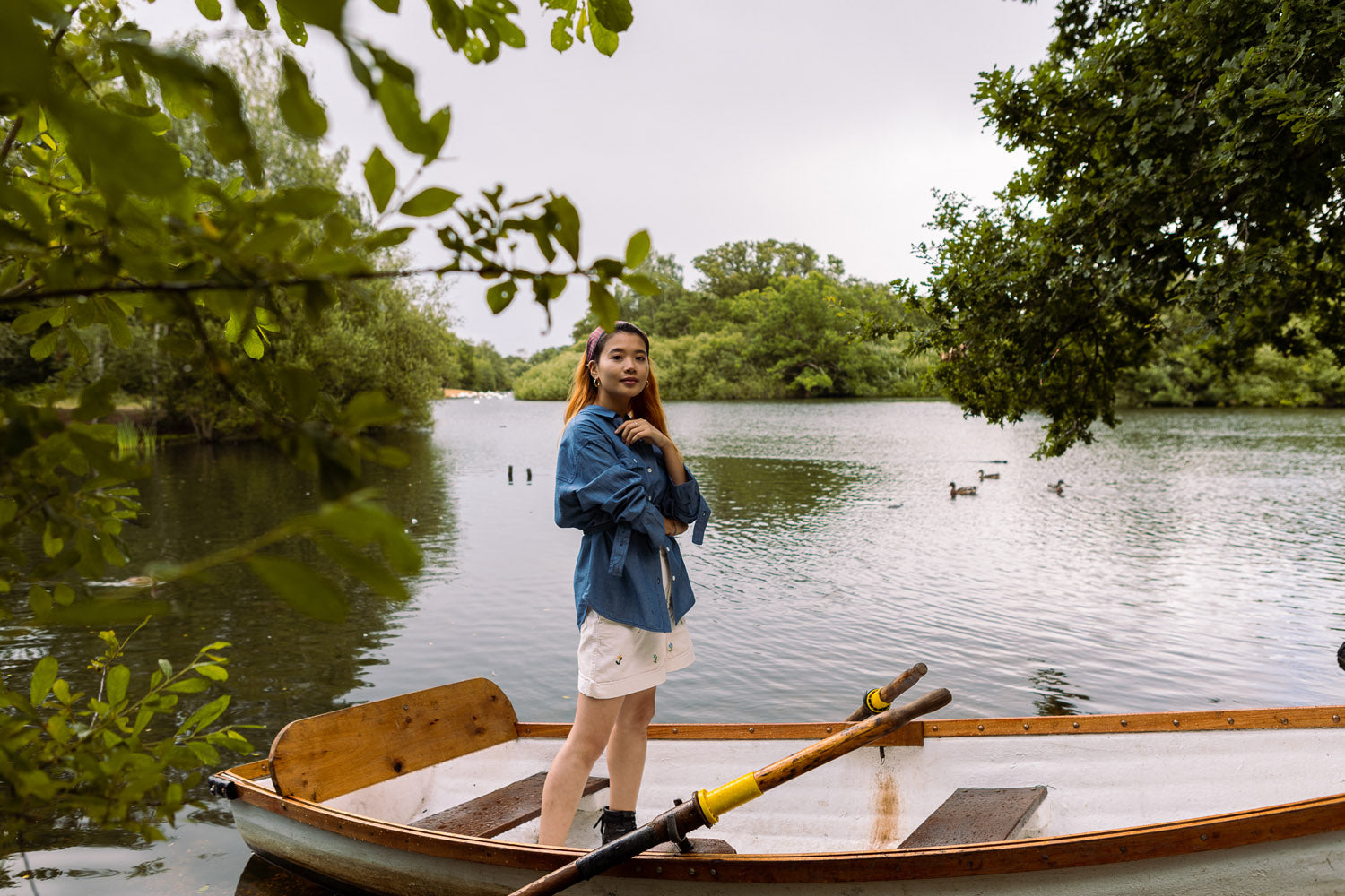 Model stands in a white row boat, with one arm resting across her waist and the other just below her chin. She wears Saywood's Japanese denim Zadie Boyfriend Shirt, with the red check Heidi Headband in her hair, and ivory cord shorts. The boat floats in a lake surrounded by trees.