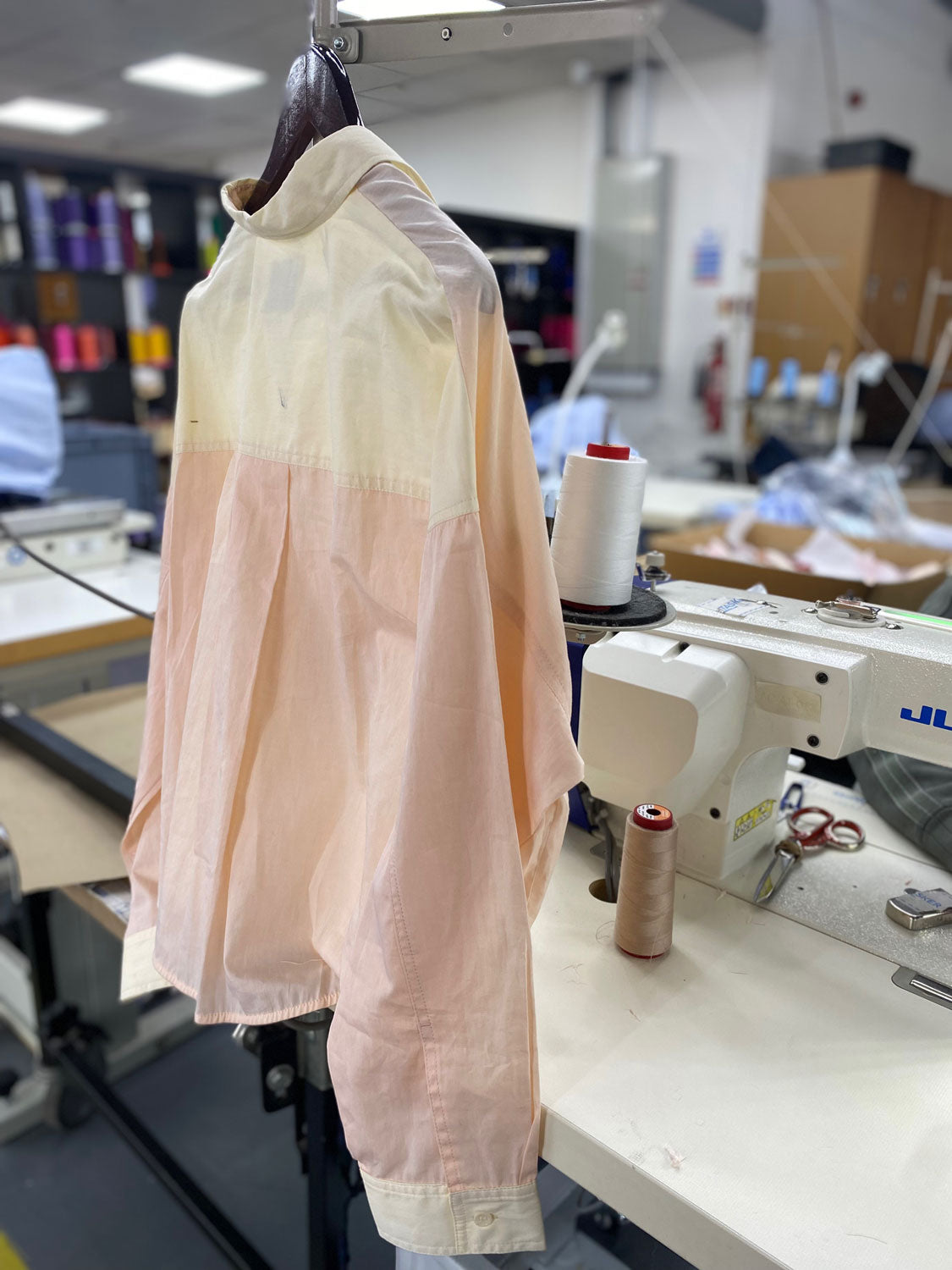 Sustainable cotton shirts made in London - Saywood's pastel orange and lemon womens Lela Shirt hangs in a factory, where sewingmachines and threads can be seen in the background