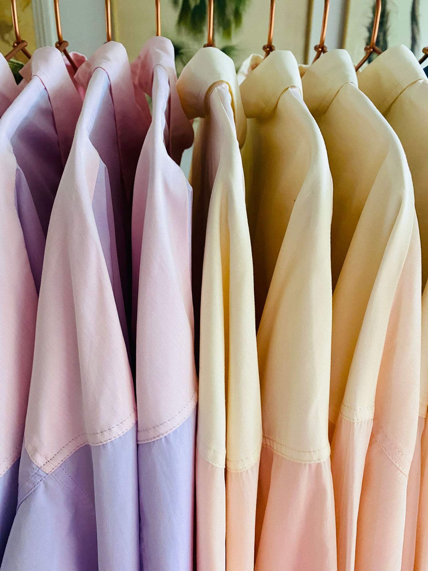 Saywood cotton shirts in lilac and pink, and yellow and orange, on rose gold hangers - a close up image showing just the shoulders, with a palm tree image vaguely seen in the background.