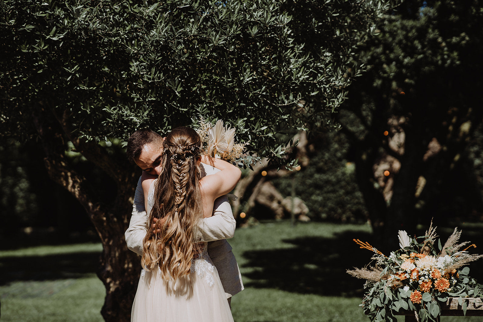 Under the trees the Bride and Groom embrace. The Bride's plaited hair is seen trailing beautifully, whilst she holds her flowers with arms wrapped around her Husband.
