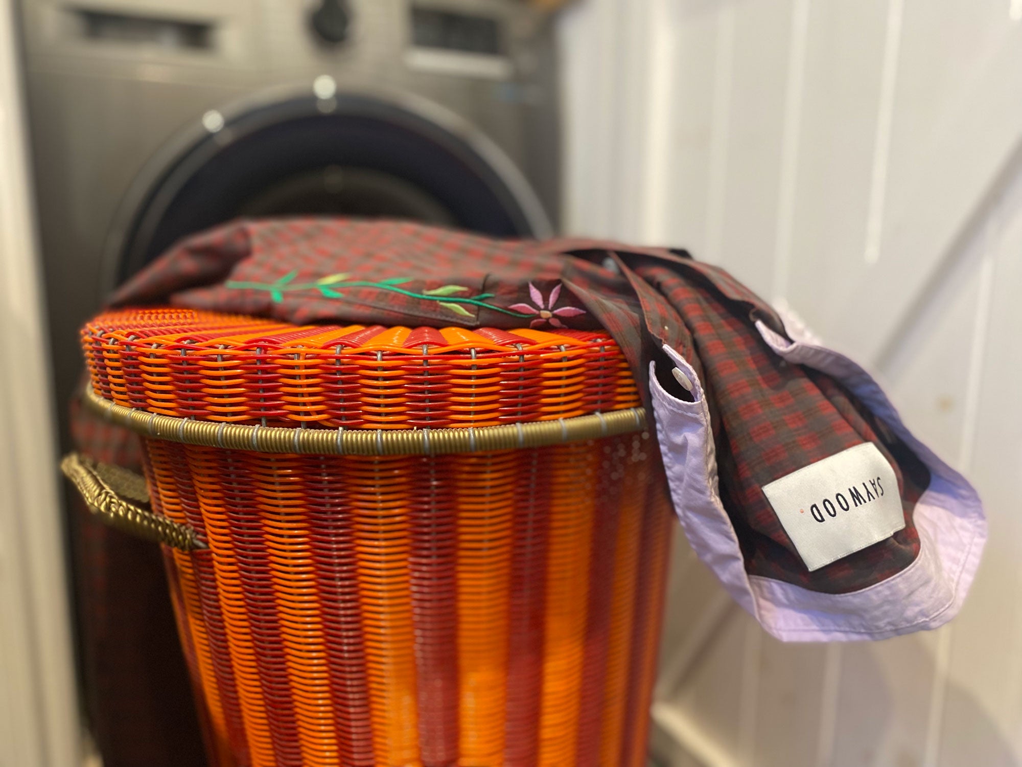 An orange and red woven laundry basket sits in front of a washing machine. On top of the basket lid lies Saywood's Etta Oversized Shirtdress in red check. The lilac collar is open and the Saywood label can be seen.