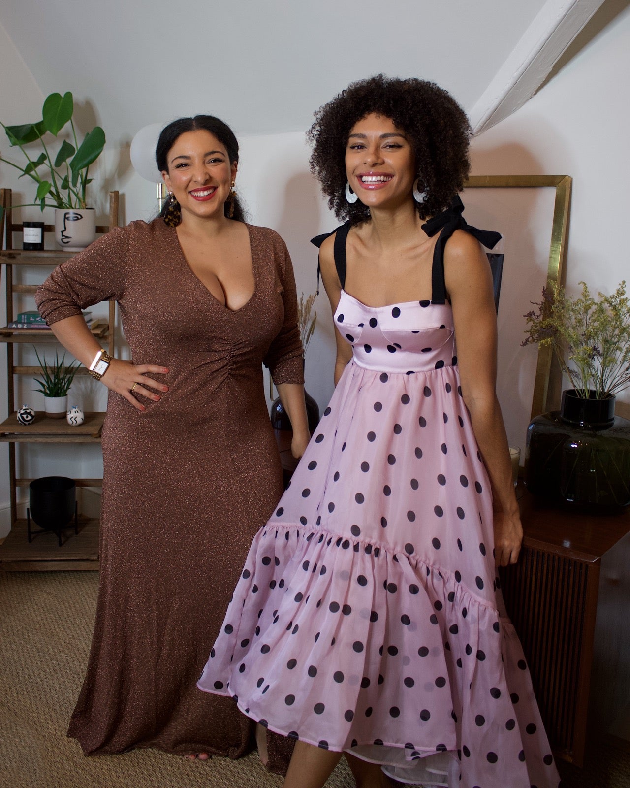 Sustainably Influenced Podcast co-founders, Bianca F Foley and Charlotte Williams, standing smiling at the camera in a living room. They both wear rented dresses; Bianca wears a chocolate sparkly v-neck dress, and Charlotte wears a lilac and black polka dot strap dress with a ruffle hem. 