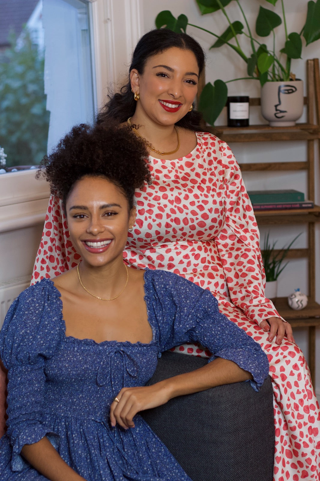 Founders of Sustainably Influenced, Charlotte Williams and Bianca Foley, sit together smiling at the camera. Charlotte wears a blue square neck dress, and Bianca wears a red and white print dress.
