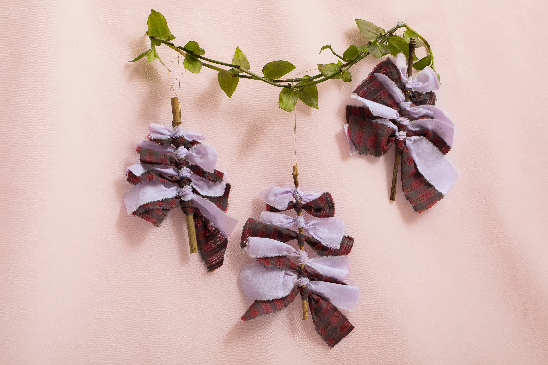 Christmas decorations: 3 Christmas Trees made from red tartan and lilac fabric offcuts tied on a bamboo stem, hanging from green micro ivy on a pink background. Sustainable decorations handmade in London