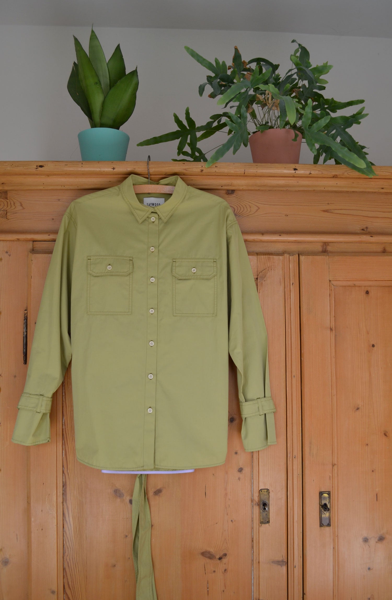 Made to last sustainable fashion. Saywood's Zadie Boyfriend Shirt in olive green, hanging on an old pine wardrobe, with two plants sitting atop the wardrobe in turquoise and pink pots.