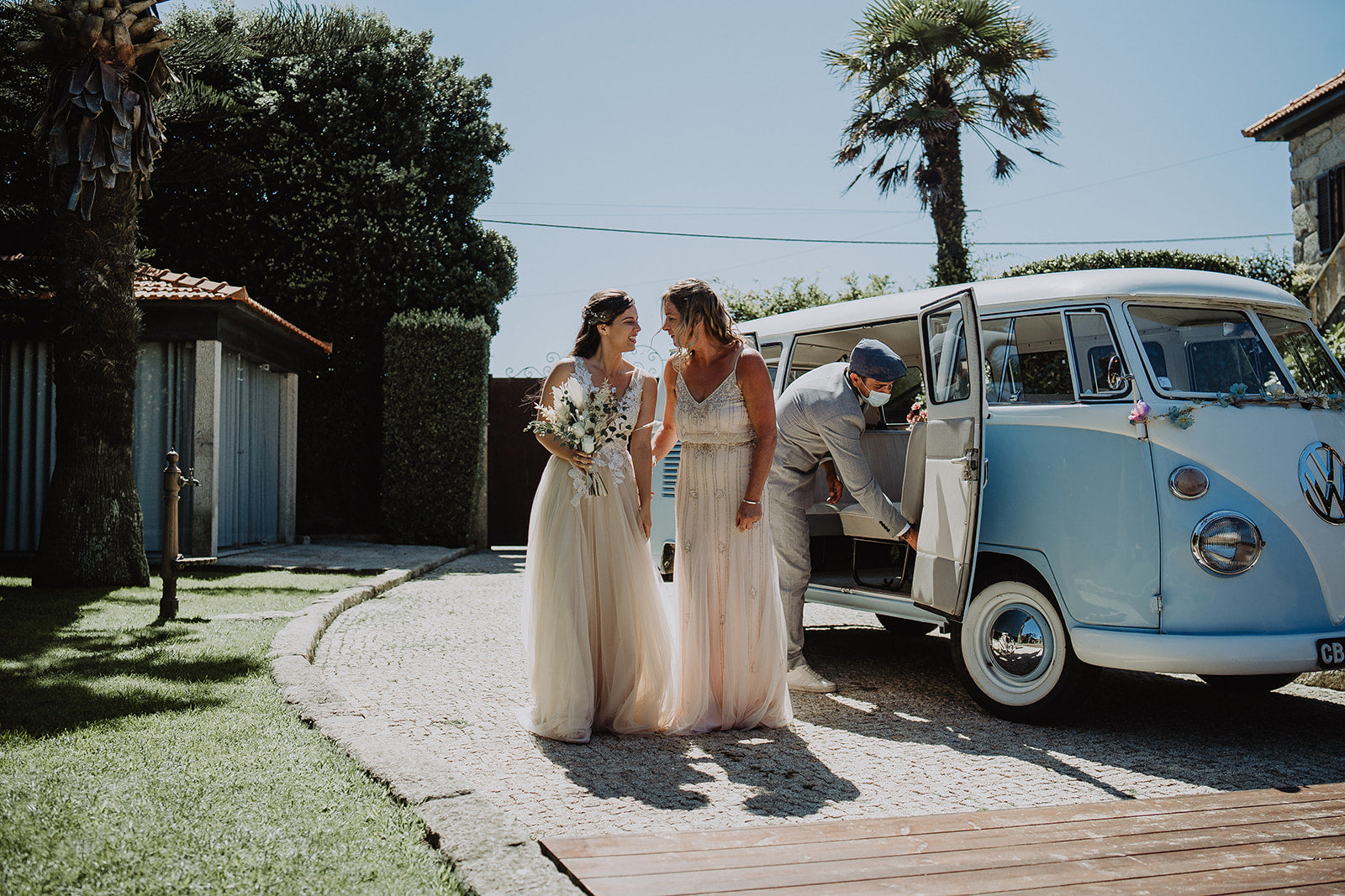 The Bride and Mother of the Bride stand together in their beautiful pre-loved dresses, with the Bride holding flowers. They stand next to a light blue VW Camper Van with a palm tree in the background.