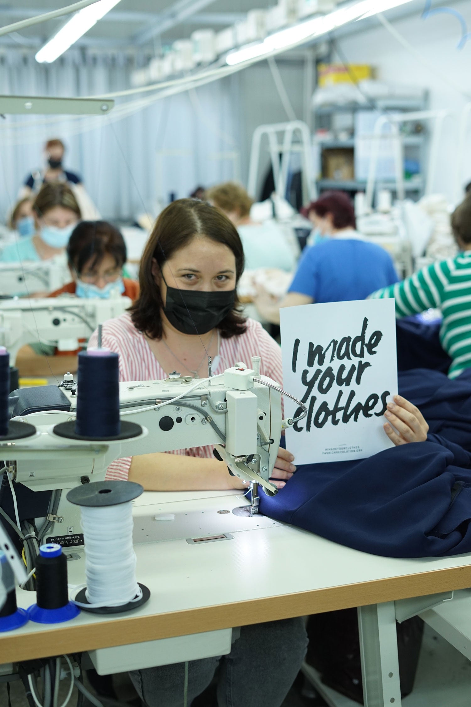 Image shows a garment factory room with a female sewing machinist sitting at her machine holding a sign with the text 'I made your clothes' for Fashion Revolution Week