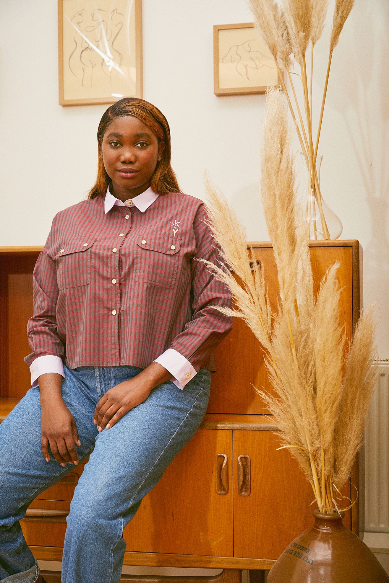 Saywood deadstock cotton Jules Utility Shirt in red check worn with jeans by model leaning against a mid century cupboard unit, with pampas grass  in a vintage vase to the right