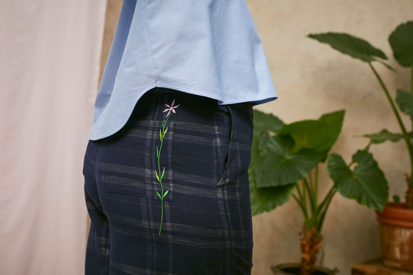 Close up side view of Saywood's Amelia check trousers, wide leg culotte shape in navy check. The lilac flower embroidery is visible on the back patch pocket. Worn with the pale blue shirt, the Marie a-line blouse. A plant and drop of pink fabric can be seen in the background.