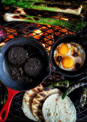 A BBQ breakfast over flames with cast iron skillets and fried eggs, black pudding, flat breads and charred leeks