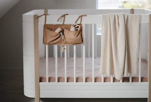 An image of a Mushie baby blanket hung over the side of a baby’s crib