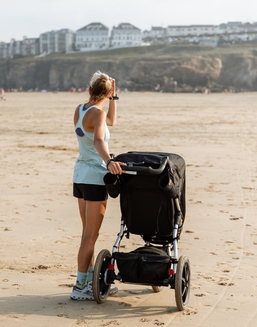 Mother on beach sand standing next to her 3 wheeled baby pram - Julian Davis | Mountain Buggy influencer