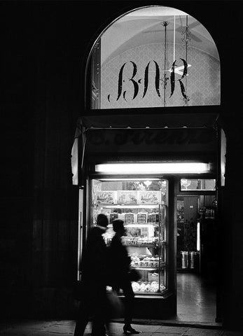 A black and white image of the outside of a bar in Florence at night with people walking by. 