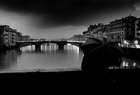 A black and white image of someone standing under an umbrella looking out to the river and some bridges in Florence.