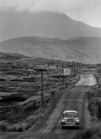 A Morris Minor travelling down a country road in Connemara with a misty mountain top in the background.