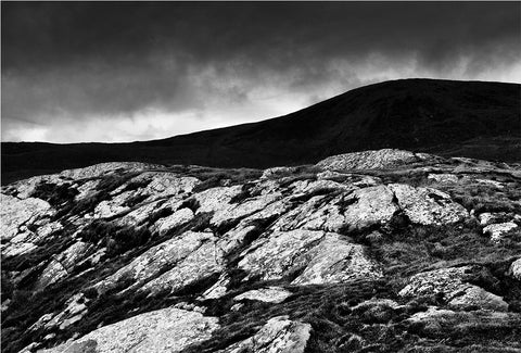 A black and white image of a rock formation in the foreground with a silhouette of a mountain peak in the background.