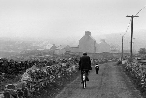 A man cycling down a stone-walled lane with a dog in front of him.