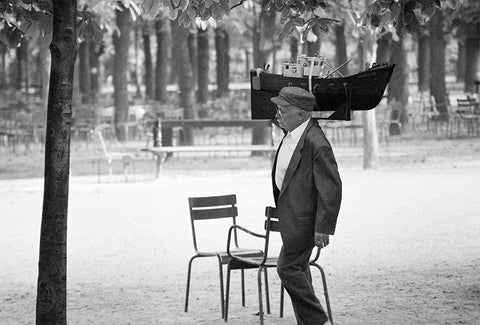 A black and white photograph of a man walking through a Parisian park holding a wooden boat on his shoulder.