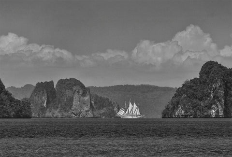 A black and white image of a large sailing boat on the water with small islands and the coastline in the background. 