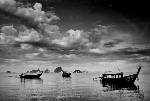 Three long-tail boats on the water with brooding clouds overhead and small islands in the background.